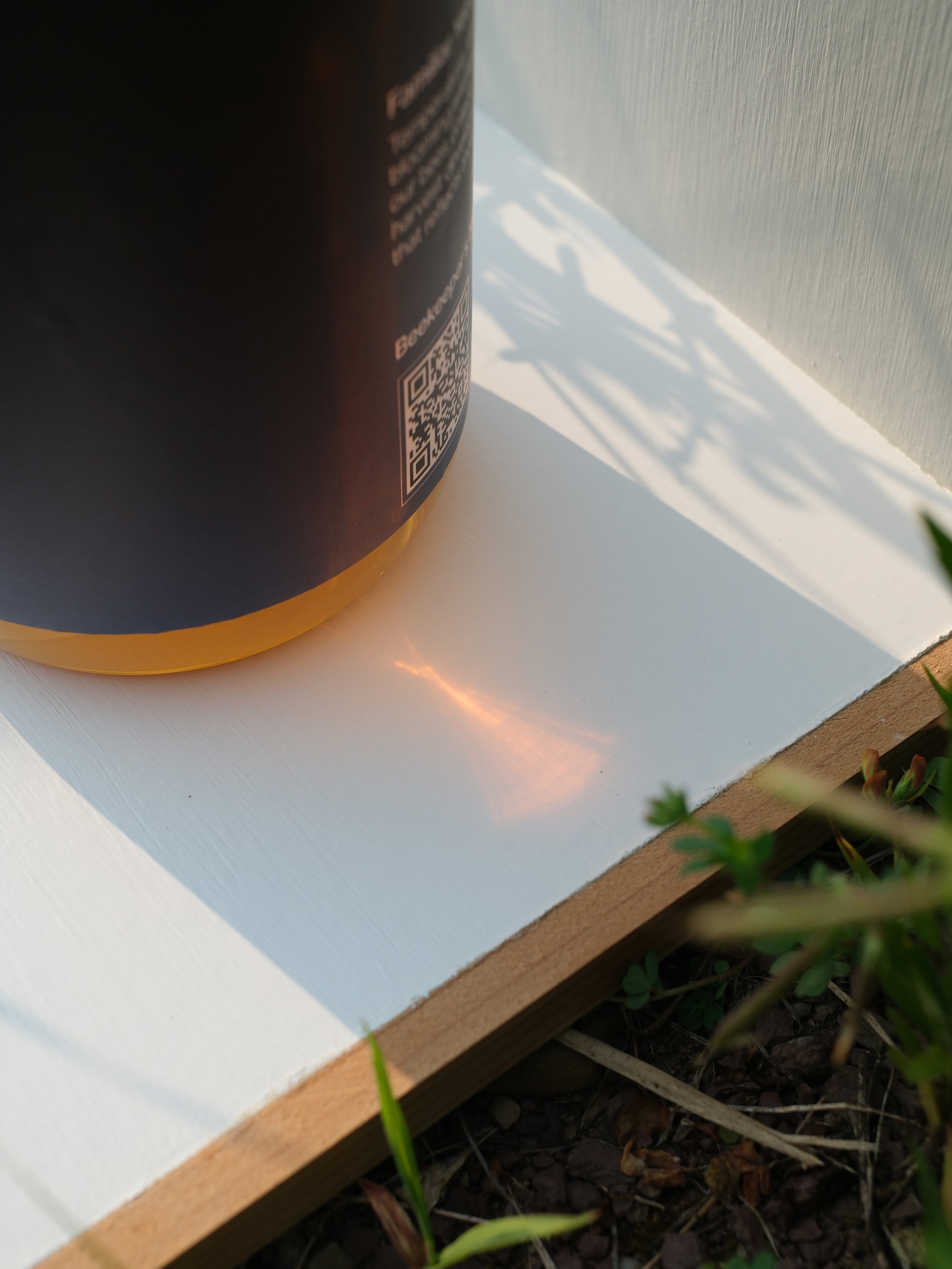 Close-up of a honey jar with a QR code on a white surface, with grass and wooden edge in the foreground.